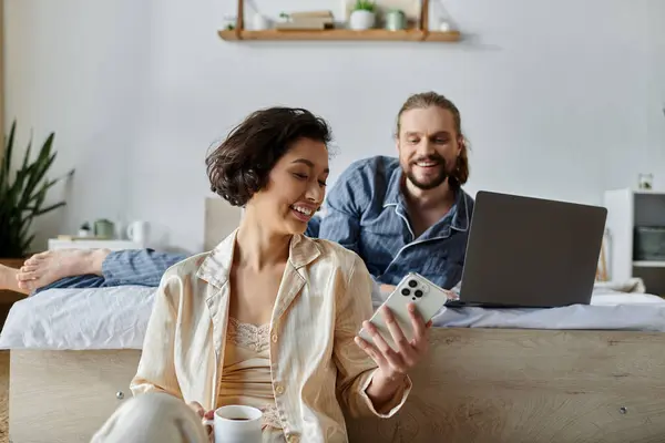 A couple relaxes on a bed, one browsing on a phone, the other using a laptop. — Stock Photo
