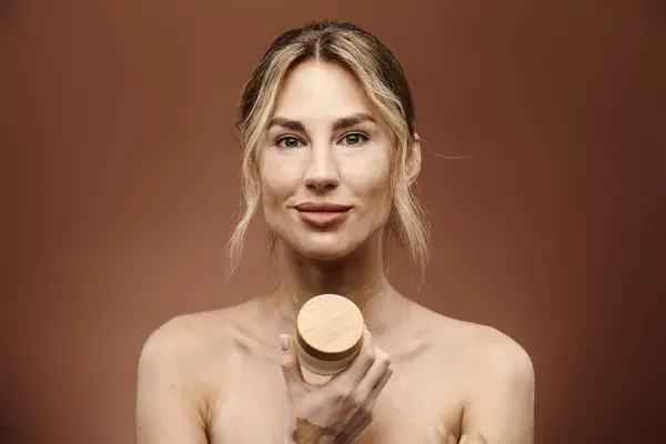 Una joven con vitiligo sostiene un frasco de crema frente a un fondo beige. Ella sonríe y mira directamente a la cámara. - foto de stock