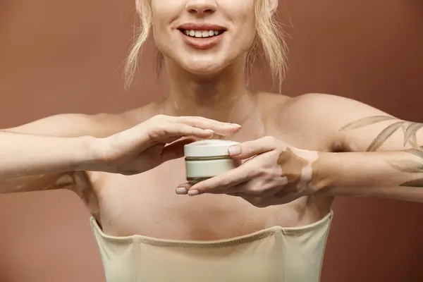 A young woman with vitiligo and a tattoo smiles while holding a jar of cream against a beige background. — Stock Photo
