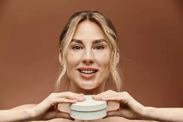 Una joven con vitiligo sonríe y sostiene un frasco de crema frente a ella sobre un fondo beige. — Stock Photo