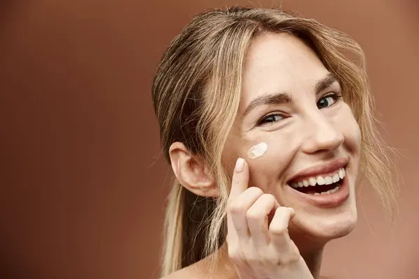 A young woman with vitiligo applies cream to her face, smiling brightly against a beige background. — Stock Photo