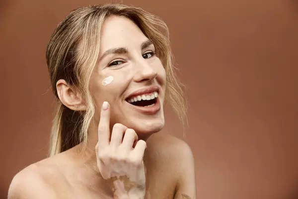A young woman with vitiligo smiles as she applies cream to her face against a beige background. — Stock Photo