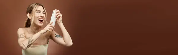 A young woman with vitiligo smiles, displaying a skin care product against a beige background. She wears a casual top and arm tattoo. — Stock Photo