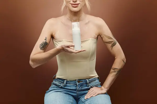 A young woman with vitiligo, wearing a tank top and jeans, holds a bottle of skincare product in front of a beige background. — Stock Photo