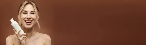 A young woman with vitiligo smiles brightly while holding a skincare product in front of a beige background. — Stock Photo