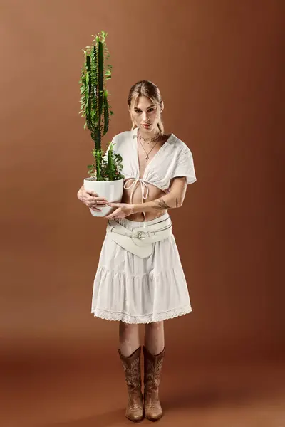 A young woman in white summer attire poses with a potted cactus against a beige background. — Stock Photo
