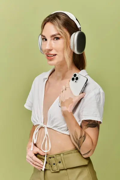 Young woman confidently poses in white outfit against green background, sporting headphones and holding phone. — Stock Photo