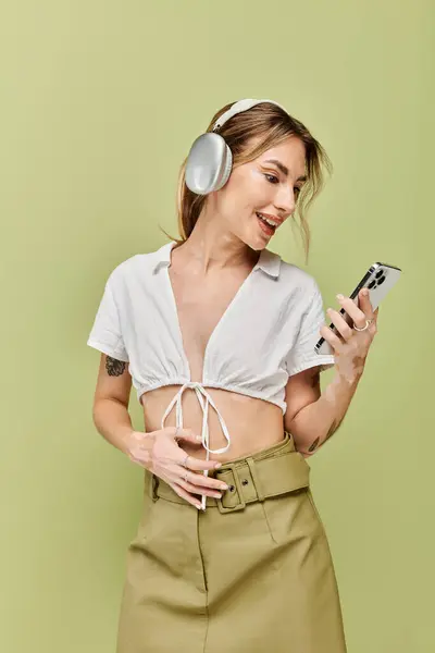 A young woman in a white top and khaki skirt poses against a green background, wearing headphones and smiling as she looks at her phone. — Stock Photo