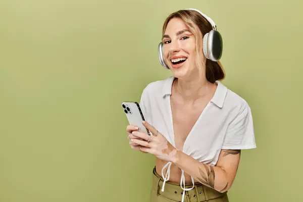 A young woman with vitiligo poses in a white summer outfit against a green background, while smiling and holding a smartphone. — Stock Photo