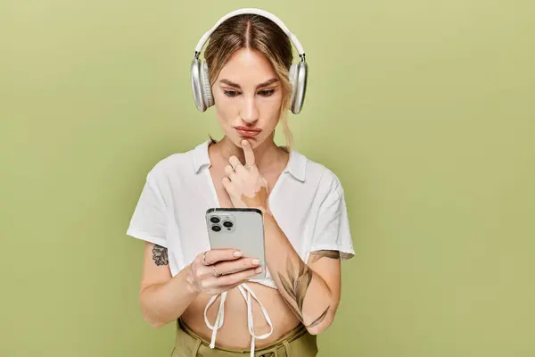 Una joven vestida con un traje blanco de verano posa sobre un fondo verde, mirando cuidadosamente su teléfono. - foto de stock