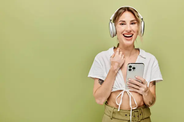 A young woman wearing a white top, khaki pants, and headphones smiles brightly while holding a smartphone. — Stock Photo