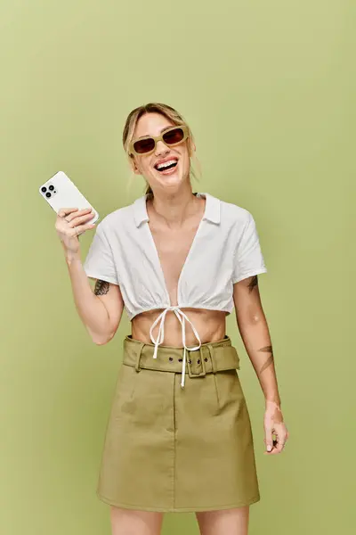 Une jeune femme avec des poses de vitiligo dans une tenue d'été blanche sur un fond vert. Elle sourit et tient un téléphone, rayonnant de confiance. — Photo de stock