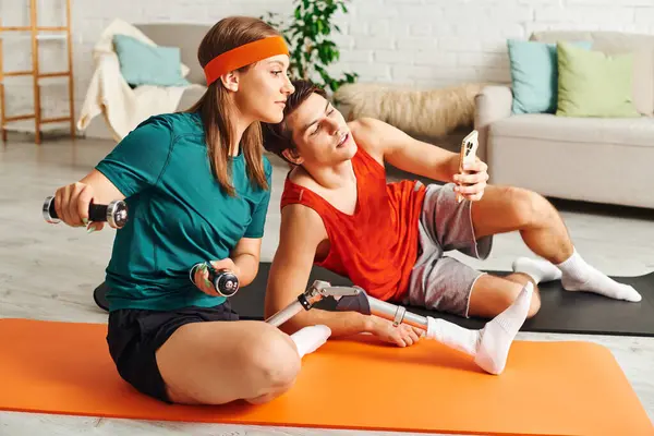 A woman with a prosthetic leg exercises with dumbbells while her boyfriend uses his phone. — Stock Photo