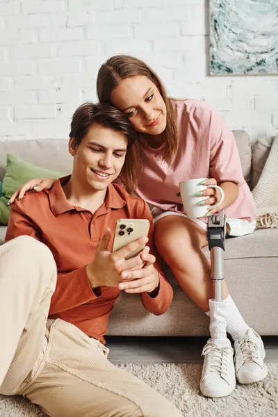 A young couple relaxes at home, with the womans prosthetic leg while her boyfriend is on his phone. — Stock Photo