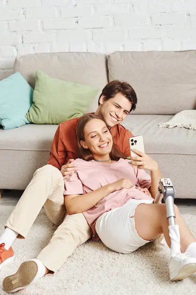 Couple bonds on rug, woman with prosthetic leg, looking at phone. — Stock Photo