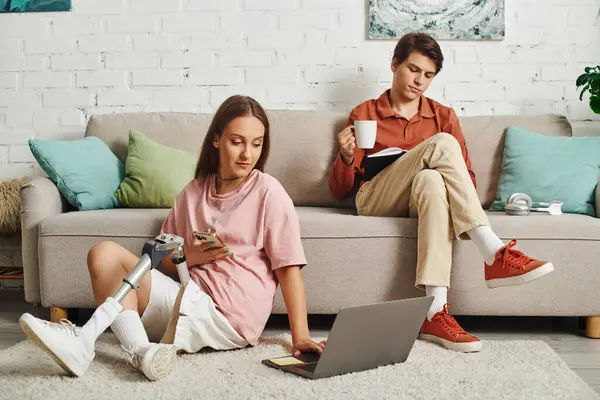 A woman with a prosthetic leg sits on the floor while her boyfriend reads a book on the couch. — Stock Photo