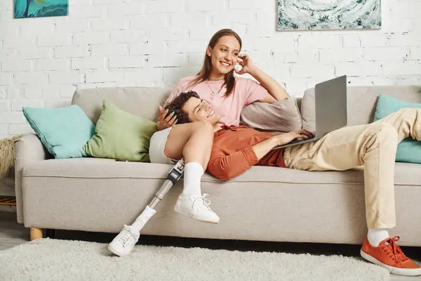 Una mujer con una pierna protésica se sienta con su novio en un sofá; usa una computadora portátil. - foto de stock