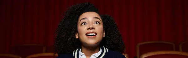 A young African American woman watches a movie in a theater, reacting with wide eyes and a surprised expression. — Stock Photo