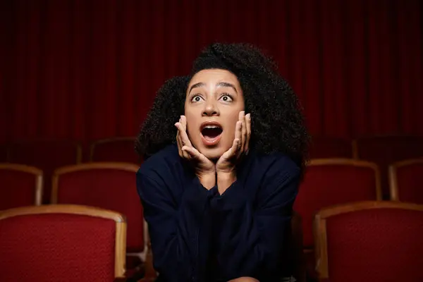 Una giovane donna afroamericana in un cinema reagisce con sorpresa, con le mani che si coprono la bocca. — Foto stock