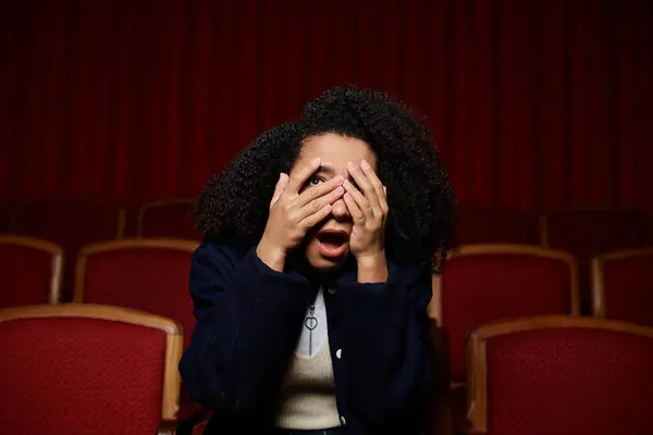 Uma jovem mulher em um cinema, cobrindo seu rosto com as mãos em uma reação dramática ao filme que joga na tela. — Fotografia de Stock