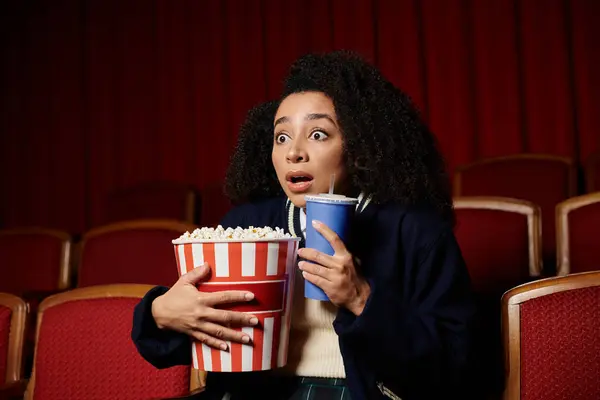 Uma jovem assiste a um filme em um teatro, segurando pipocas e uma bebida enquanto reage a uma cena surpreendente. — Fotografia de Stock