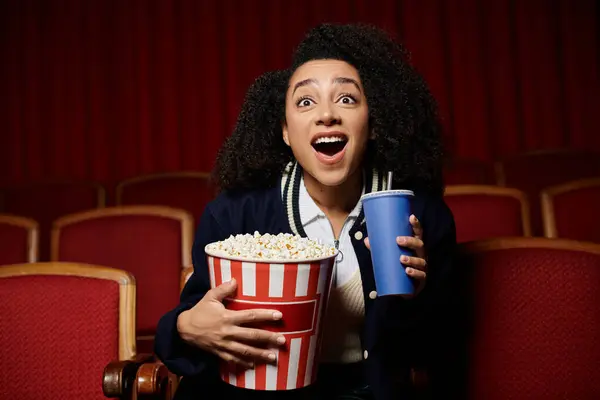 Eine junge Frau im Kino sieht sich einen Film an, hält einen Eimer Popcorn und ein Getränk in der Hand und reagiert mit Aufregung und Überraschung. — Stockfoto
