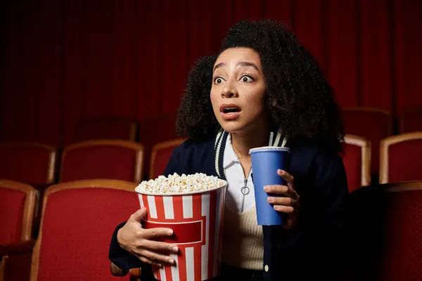 A young woman with a look of surprise sits in a movie theater, holding popcorn and a drink. — Stock Photo