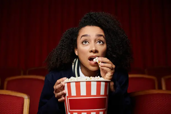 A young woman in a movie theatre watches the screen with a surprised expression, eating popcorn. — Stock Photo