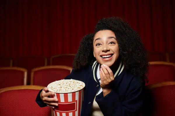 Young Black woman in chic attire, enjoying movie at cinema with popcorn in hand, laughing and engrossed in the film. — Stock Photo