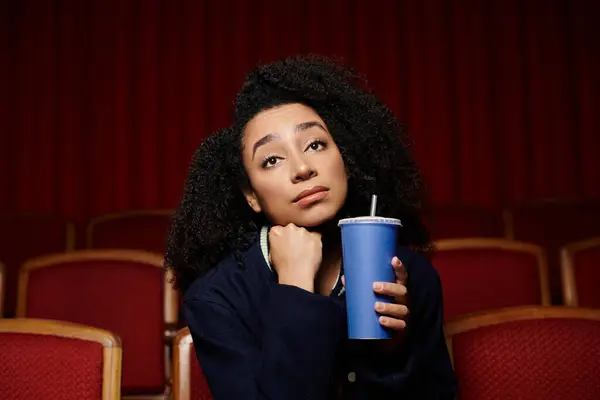 A young African American woman sits in a theater seat, thoughtfully watching a movie, with a beverage in hand. — Stock Photo