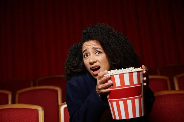 Uma jovem afro-americana reage de surpresa enquanto assiste a um filme no cinema, agarrando seu balde de pipoca. — Fotografia de Stock