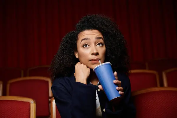 Une jeune femme aux cheveux bouclés s'assoit dans un siège de théâtre, tenant un verre et regardant le film. — Photo de stock