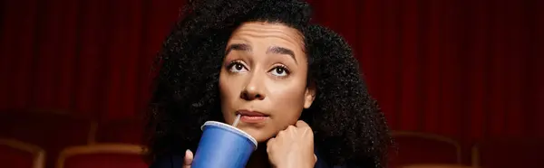 A young woman with curly hair sits in a theater seat, gazing at the screen with a thoughtful expression, taking a sip from her drink. — Stock Photo