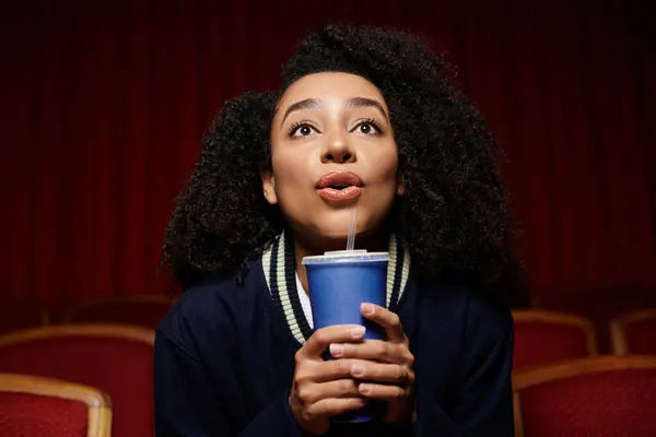A young African American woman sits in a movie theater, looking up at the screen with a surprised expression, holding a drink. — Stock Photo