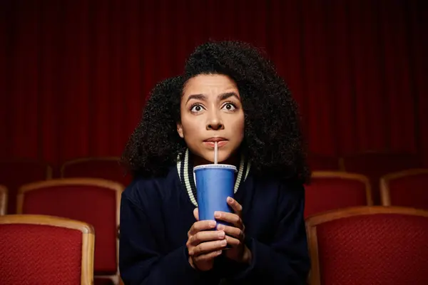 Uma jovem mulher em roupas casuais senta-se em um assento de teatro, segurando uma xícara e olhando para cima de surpresa, aparentemente absorvida em um filme. — Fotografia de Stock