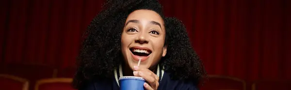 Una giovane donna afroamericana dai capelli ricci si siede in un cinema, ridendo e bevendo da una tazza. — Foto stock