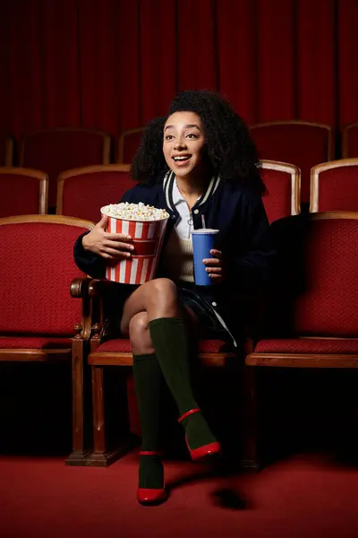 A young woman watches a movie, her expression filled with excitement as she enjoys a bucket of popcorn and a drink. — Stock Photo