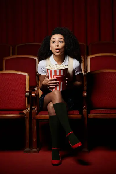 A young woman sits in a cinema seat, eyes wide with surprise as she watches the movie. — Stock Photo