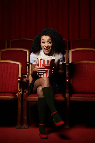A young woman in a theater seat excitedly watches a movie, holding a bucket of popcorn. — Stock Photo