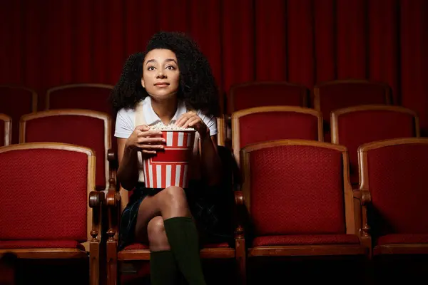 Uma mulher afro-americana em um assento de filme, assistindo o filme com espanto, segurando um balde de pipocas. — Fotografia de Stock