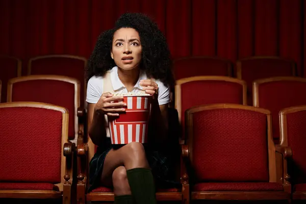 Uma jovem se senta em um assento de cinema, segurando seu balde de pipoca e olhando chocado para a tela. — Fotografia de Stock