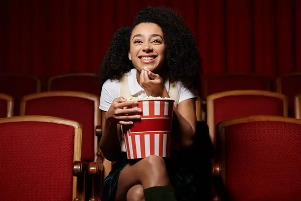 A young woman with curly hair sits in a theater seat, happily enjoying a movie and a bucket of popcorn. — Stock Photo