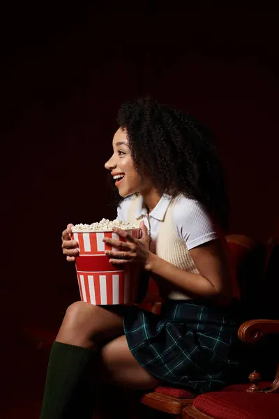 A young woman in stylish casual clothing watches a movie in a cinema, laughing and holding a bucket of popcorn. — Stock Photo