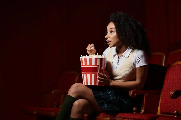 A young woman captivated by the film on screen, sits in a theater seat with a bucket of popcorn, eagerly watching. — Stock Photo