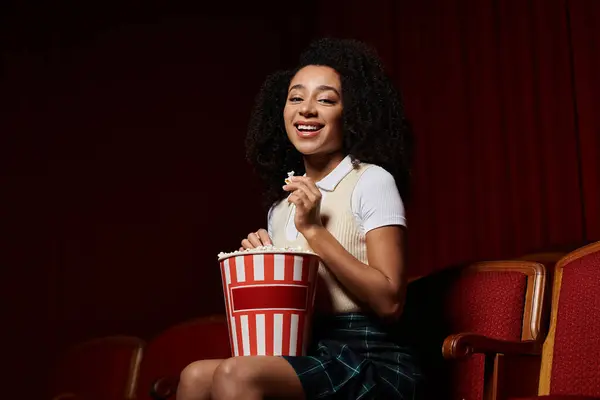 A young African American girl sits in a cinema seat, enjoying a movie with a big smile and a bucket of popcorn. — Stock Photo