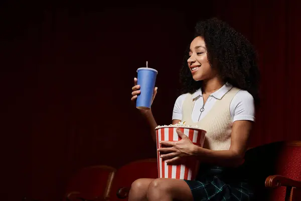 Una giovane donna con i capelli ricci si siede in un cinema, reggendo un drink e popcorn, sorridendo e godendo il film. — Foto stock
