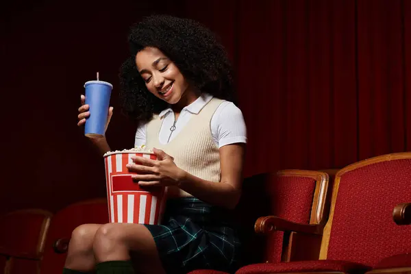 Jeune femme noire en tenue décontractée aime le film avec pop-corn et boire dans le siège du théâtre, souriant et absorbé dans le film. — Stock Photo