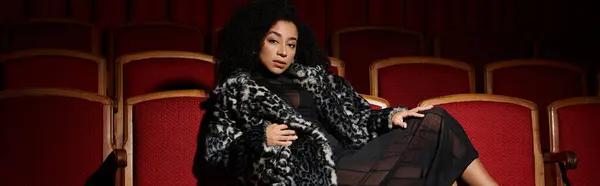 An elegant African American woman in a trendy coat sits in a theater seat, watching a movie with a focused expression. — Stock Photo