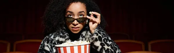A fashionable African American woman enjoys a movie, popcorn in hand, while sporting a trendy leopard print coat. — Stock Photo