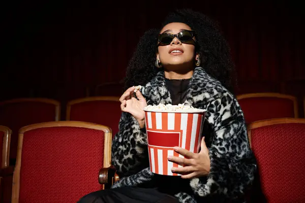 A fashionable woman wearing a leopard print coat watches a movie in a darkened cinema, enjoying popcorn. — Stock Photo